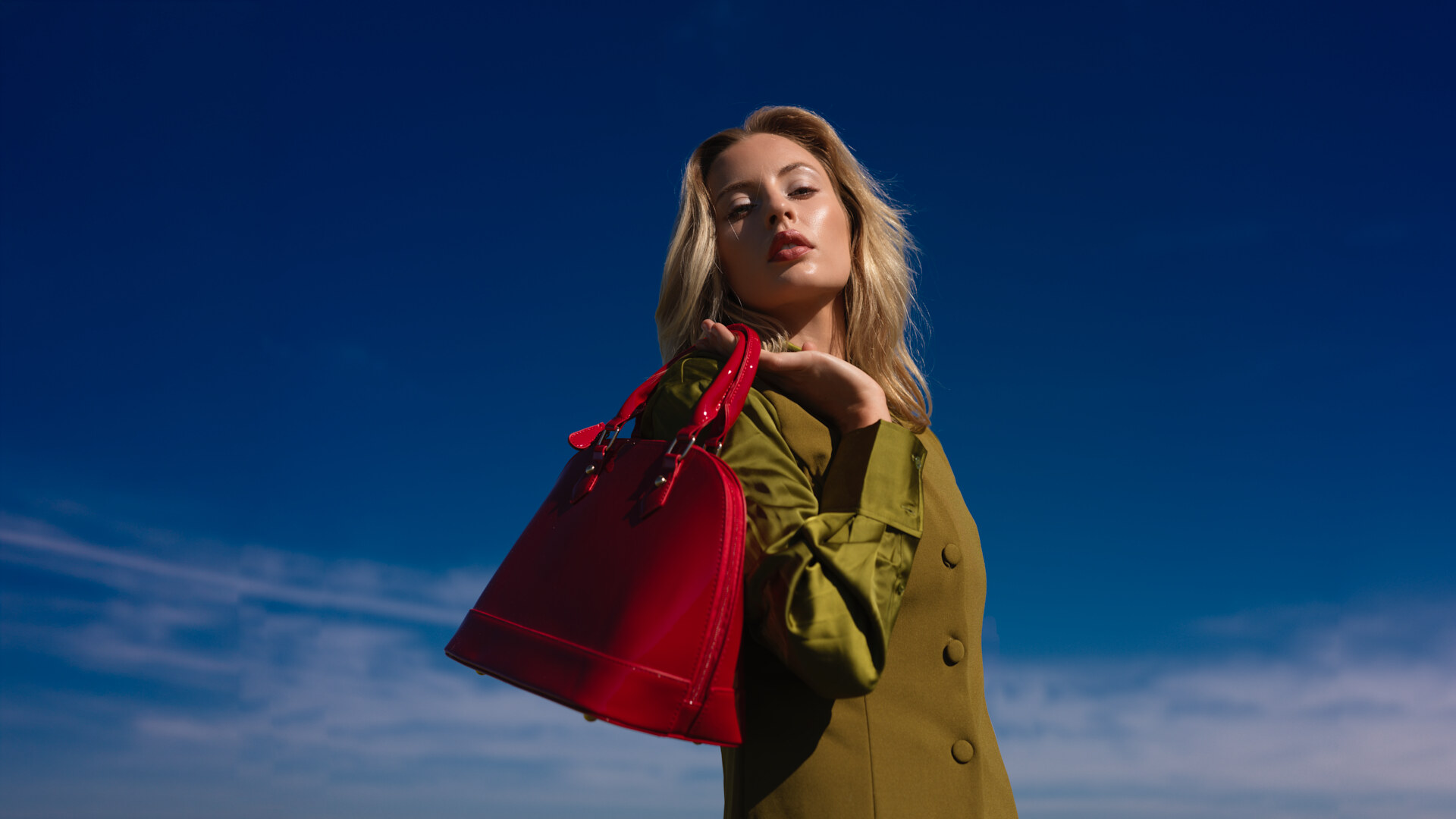 Fashion accessories editorial of a model holding a red patent leather handbag against a clear blue sky, styled in an olive green utility coat in bright natural light. Jason Guffy Photography.
