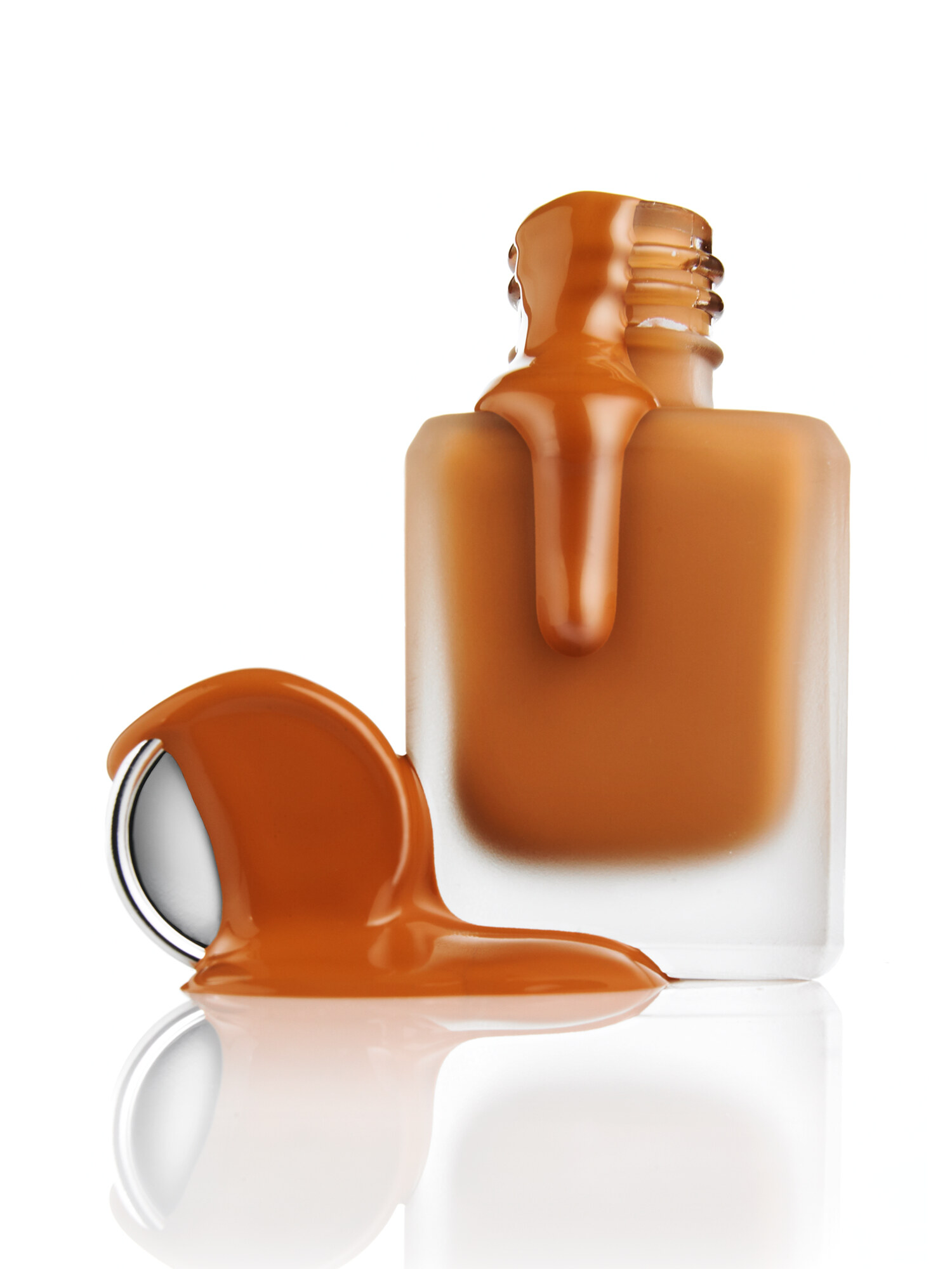 Editorial product still life of liquid foundation spilling from a frosted glass bottle with detached cap on a glossy reflective surface. Beauty advertising product photography by Jason Guffy.