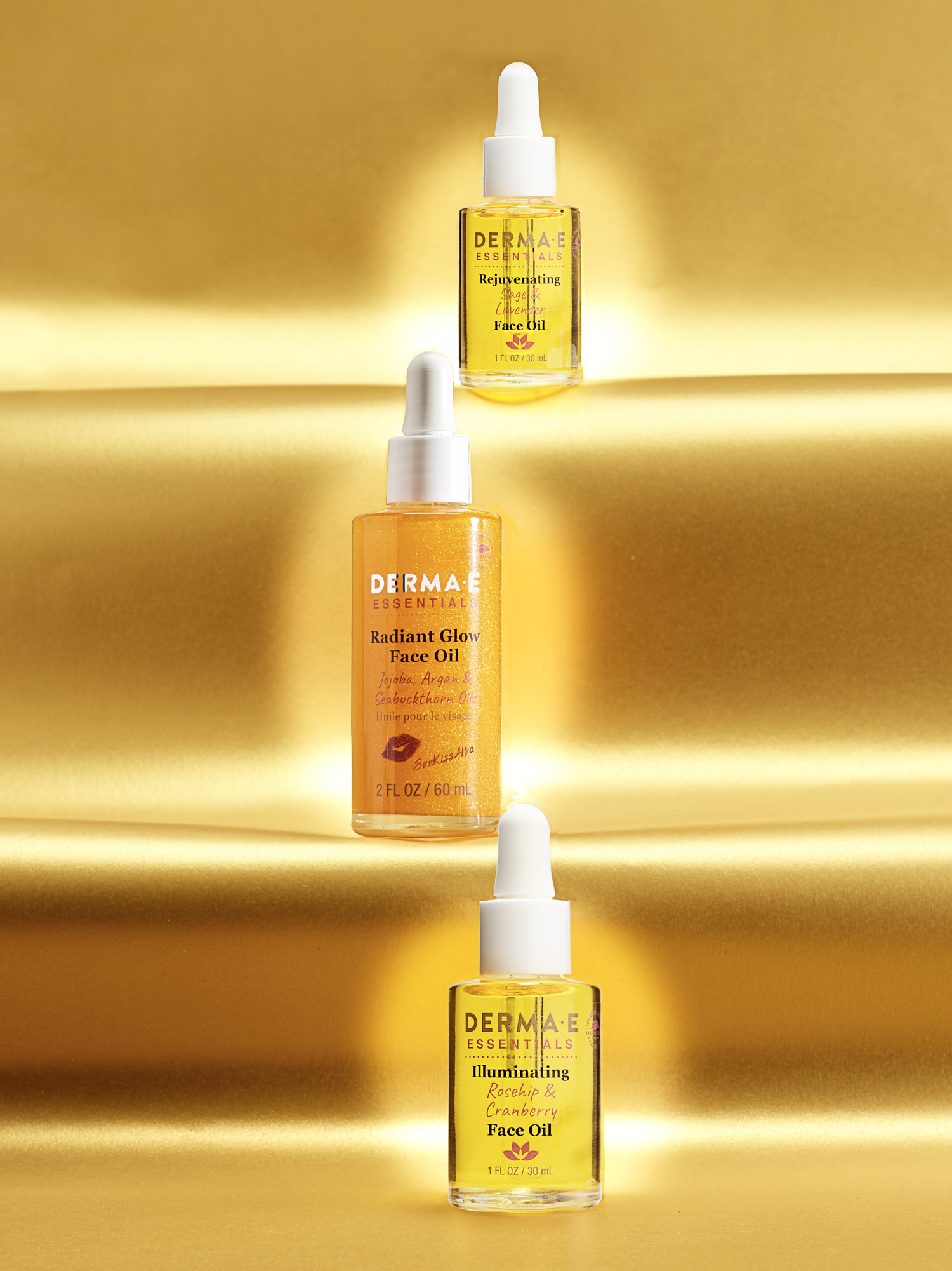 Derma E Essentials face oil trio, Radiant Glow, Rejuvenating Sage and Lavender, and Illuminating Rosehip and Cranberry, suspended against a gold satin backdrop. Skincare product photography by Jason Guffy.
