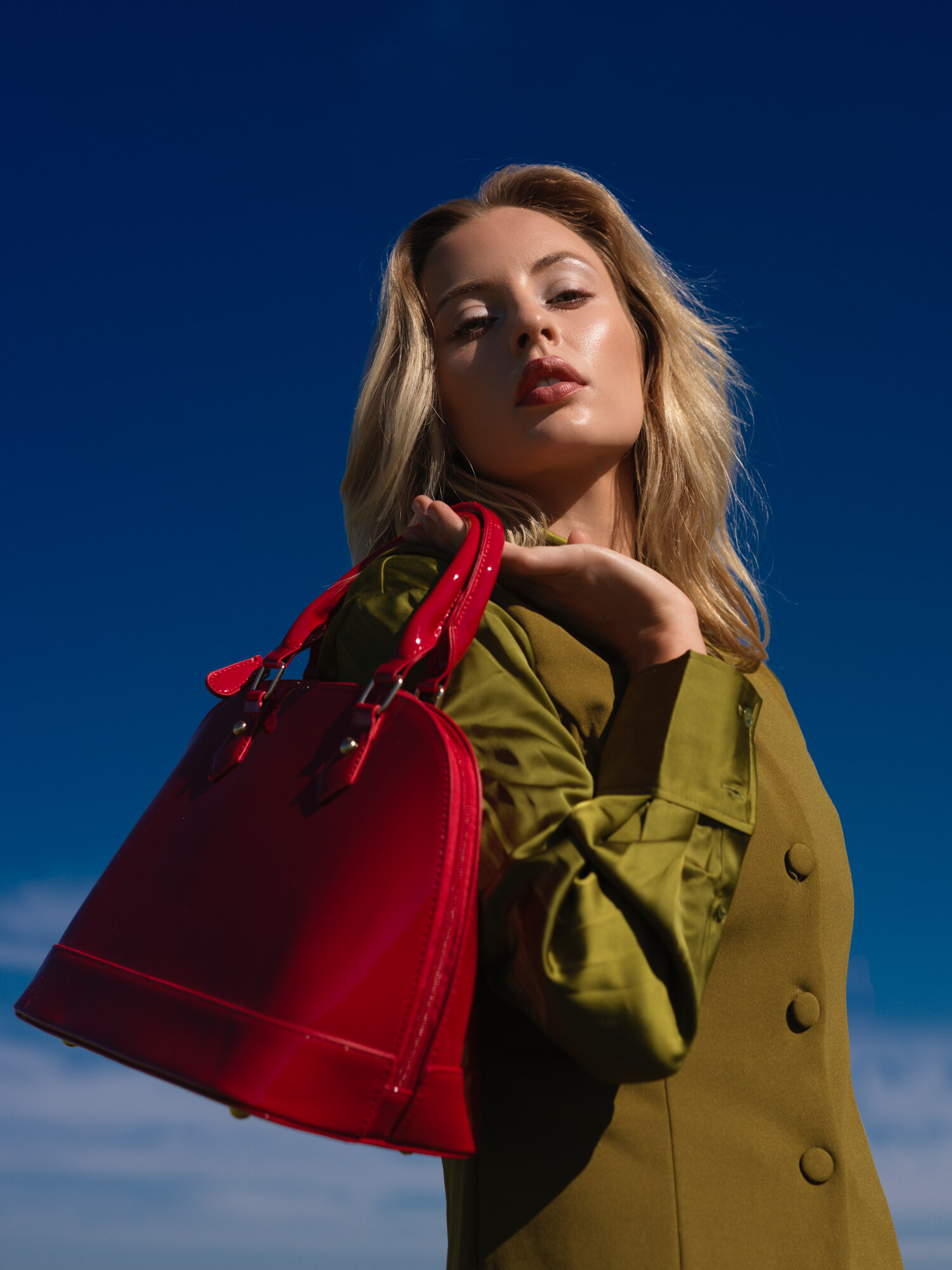 Fashion lifestyle editorial holding a red patent handbag in a grass field, olive knit dress in natural daylight. Fashion photography by Jason Guffy.