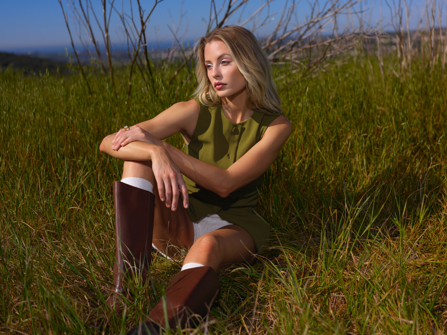 Fashion lifestyle editorial seated in a coastal grass field, olive knit dress in soft natural light. Fashion photography by Jason Guffy.