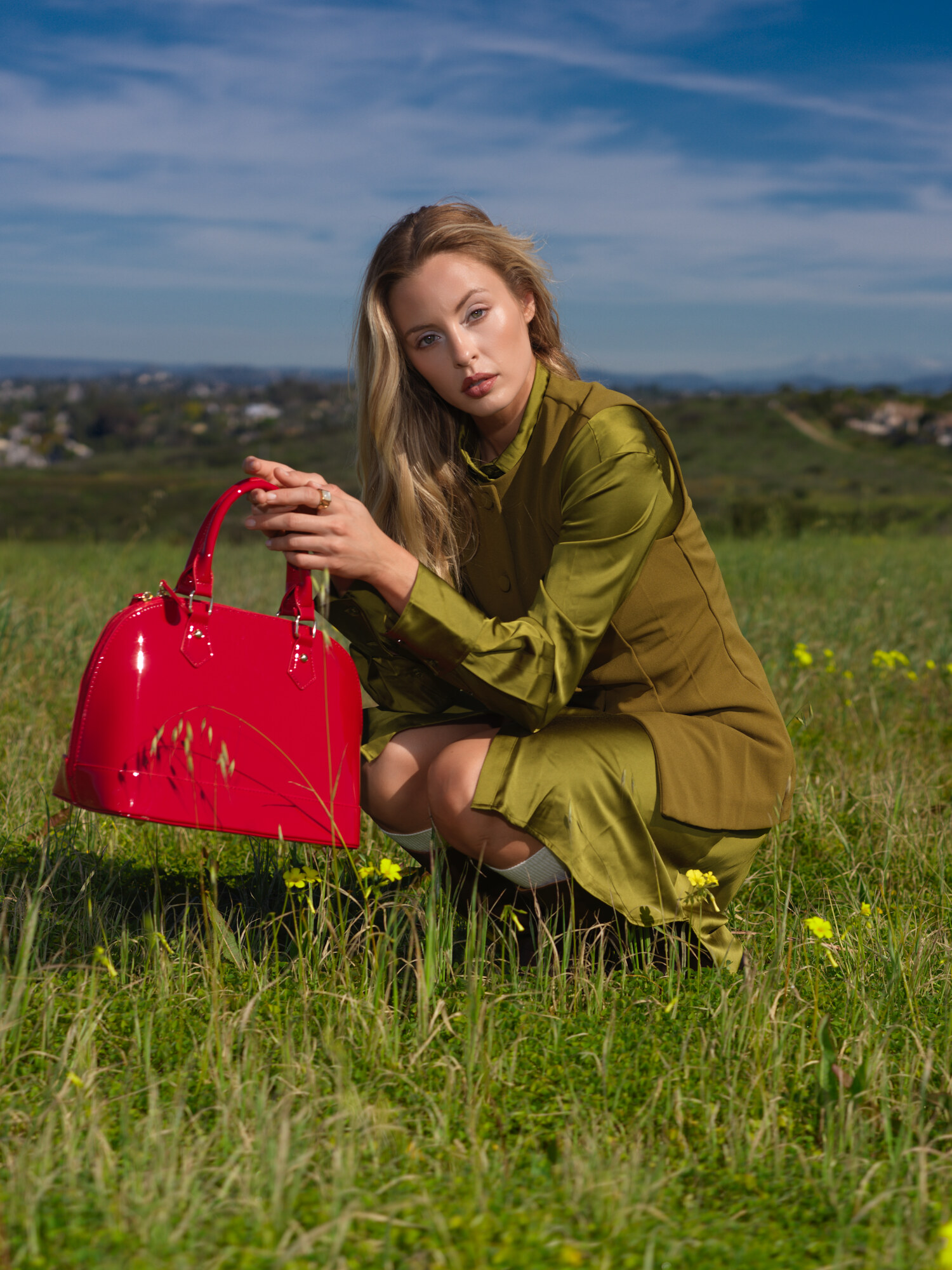 Fashion lifestyle editorial crouched among wildflowers with a red patent handbag, olive knit dress. Fashion photography by Jason Guffy.